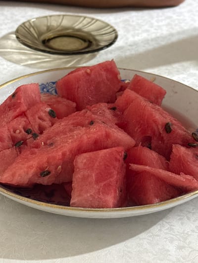 A close-up, overhead shot captures a white ceramic bowl filled with cut pieces of red watermelon. The watermelon slices are irregular in shape, with visible black seeds scattered throughout. The bowl has a decorative blue pattern on its inner rim and a thin gold trim. It rests on a white tablecloth with a subtle floral embossed pattern. To the left of the bowl, a small, dark brown glass dish casts a shadow on the tablecloth. The background shows a blurred human torso and arm. This image was taken indoors, likely in a domestic setting, in the city of Talas, Kyrgyzstan.