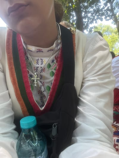 The scene depicts the upper body of a person, observed from a slightly elevated perspective, outdoors in Bolyarovo, Bulgaria. The central figure wears a white, long-sleeved garment, likely a traditional shirt or dress, featuring a high collar with intricate, multi-colored embroidery in geometric patterns, visible on the chest and collar area. The V-neck opening is trimmed with a band of vertical stripes in colors including orange, white, dark green, and red. A silver-colored chain necklace with a small metallic crucifix pendant rests on the embroidered fabric. A black strap, possibly from a shoulder bag or fanny pack, crosses over the upper left chest. Near the upper left shoulder, a spherical, gold-colored adornment, potentially a decorative button or brooch, is visible. The person's lower face, including the lips and chin, is partially visible, oriented slightly away from the camera. In the lower left foreground, a blue plastic water bottle with a turquoise screw-cap is positioned. To the far right, a portion of another person's clothing, also white with traditional red, green, and blue embroidery, is discernible. The background consists of blurred green foliage and tree canopies against a bright sky, indicating a sunny outdoor environment.