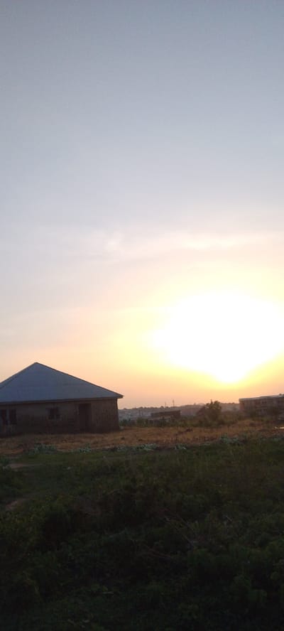The image shows a landscape at sunrise in Ibo, Nigeria. A building with a blue-grey roof stands to the left. In the distance, there are more buildings and structures. The ground is covered with vegetation. The sky is light, transitioning from yellow-orange near the sun to light blue.