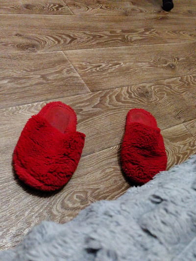 The image shows a pair of red fuzzy slippers on a wooden floor. The slippers are positioned side-by-side, with the left slipper slightly more prominent. A blurred gray fuzzy blanket is visible in the foreground, partially obscuring the slippers and floor. The wooden floor has a prominent grain pattern. There is no indication of the city of Okhtyrka, Ukraine, or any other specific location. The scene depicts a domestic indoor environment.