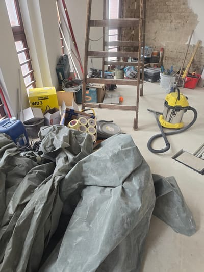 The image displays an indoor construction or renovation site located in Nyíregyháza, Hungary. No individuals are present in the scene.
The foreground is dominated by a large, rumpled gray protective tarp, covering a significant area of the light-colored, smooth floor. To the left of the tarp, a blue plastic jerry can, partially labeled "Primer G," rests on the floor, surrounded by coiled electrical cables and hoses.
Centrally, a tall, aged wooden ladder with multiple rungs stands upright. To the left of the ladder's base, a yellow Kärcher WD3 box is stacked on other cardboard boxes, next to a black bucket. A black Makita tool case, more coiled cables, and various smaller boxes are situated around the base of the ladder. To the right of the ladder's base, an open cardboard box holds several rolls of yellow masking tape, adjacent to a gray plastic lid and another container on the floor.
In the right mid-ground, a yellow and silver Kärcher wet/dry vacuum cleaner is positioned on the floor, its black hose extended towards a rectangular cut-out in the floor.
The background left features two tall, narrow windows with brown frames, allowing natural light into the room. The walls between and extending from these windows are covered with fresh, white plaster or paint. The background right wall is partially stripped, exposing raw brickwork and remnants of old plaster, indicating ongoing renovation. Leaning against this wall are a shovel, a red and silver spirit level, and various buckets (one white, one red) filled with tools or materials, alongside stacked boxes.
The floor throughout the visible area is a light, uniform surface, consistent with a concrete or screed finish typical of a renovation project. The overall environment is that of an active construction zone, characterized by scattered tools, building materials, and partially completed structural elements.