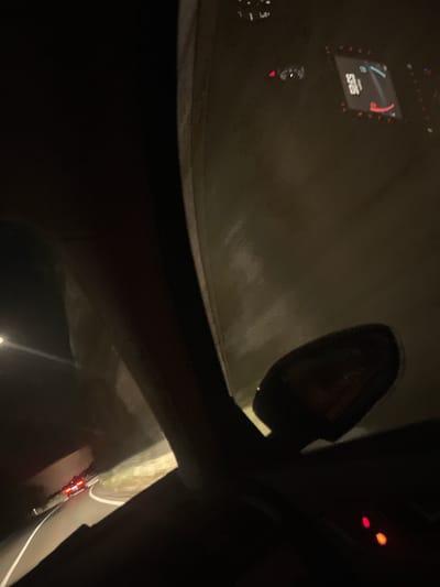 The image is taken from inside a vehicle, looking out through the windshield and side window at night. The view captures a dark, winding road, likely a mountain pass, with some vegetation and rocks visible along the side. In the distance, another vehicle with its taillights illuminated is traveling ahead on the road. The interior of the car is dark, with a portion of the dashboard and a side mirror visible. A digital display on the dashboard shows some readouts, including what appears to be a speedometer. There are no people clearly visible in the image. The overall impression is of a night drive on a rural road, possibly in the mountainous region of Golubovići, Montenegro, given the context provided.
