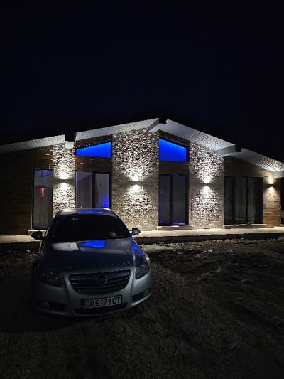 At night in Dragushinovo, Bulgaria, a silver Opel Insignia sedan is parked facing the camera on an unpaved, dark, and uneven dirt or gravel surface. The vehicle bears Bulgarian license plate "CO 5173 CT." Behind the car, a modern single-story house with a complex, multi-angled roofline is illuminated. The house features a facade of light brown brick and textured stone cladding. Several downward-facing spotlights cast white light onto the stone. Two prominent, angled windows on the front of the house glow with a distinct blue light from within, which reflects on the hood of the parked car. Multiple dark-framed glass doors or windows are visible across the facade, set above a light-colored foundation or threshold. The sky above is dark. No individuals or other objects are present in the immediate vicinity.