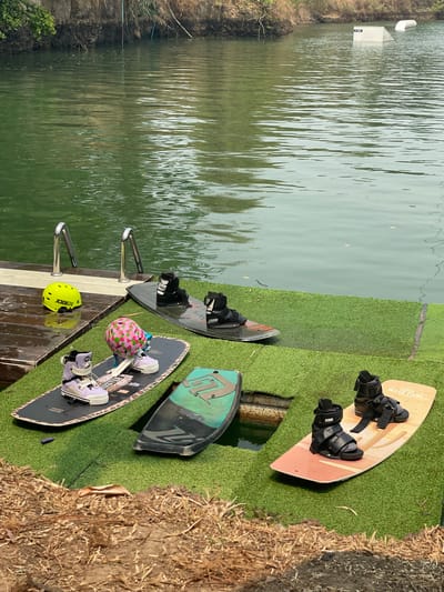 The scene depicts a waterfront area at a wake park or similar recreational facility, likely located in Tambon Nam Phrae, Thailand. The water is calm, with reflections visible on the surface. Several wakeboards are laid out on artificial turf next to the water. These include a light brown wakeboard with black bindings, a black wakeboard with black bindings, a green and black wakeboard, and a dark wakeboard with purple boots and a helmet with a pink floral design. A yellow helmet rests on a wooden platform near the water's edge. Two stainless steel handrails are visible, presumably for entering or exiting the water. The surrounding environment includes vegetation and what appears to be dry grass or straw along the edge of the artificial turf. A structure is visible in the distance across the water. There are no people present in the scene.