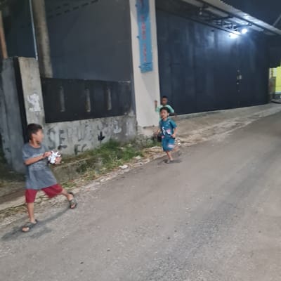 Three young boys are running down a paved street at night in Kecamatan Tamansari, Indonesia. The boys are wearing casual clothing. One boy is carrying a small white bag in his right hand. In the background, there is a building with a closed door.  A streetlight illuminates the scene. The building has faded writing on the wall. There is vegetation growing along the side of the street.  The boys appear to be playing a game together.  The location is a residential area in the city.