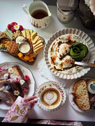 This overhead shot depicts a meticulously arranged breakfast or brunch spread laid out on a clean white surface, likely in Ros Na Rí, Ireland. The scene suggests a leisurely, indulgent meal.

In the center, a large white scalloped plate holds two perfectly poached eggs, a spiral of sliced avocado, and is artfully drizzled with a dark sauce, sprinkled with paprika and dill. A "ZWILLING" branded serrated knife is partially embedded in this dish. To the upper right, a smaller plate features two slices of artisan bread and a container of "BEURRE D'ISIGNY" butter. Below this, a delicate teacup, held by a hand with a floral-patterned sleeve, displays intricate latte art on its surface, resting on a matching floral saucer.

In the lower left, a charcuterie board is laden with a variety of cheeses (sliced and cubed), several types of crackers, walnuts, and fresh raspberries. A small jar on this board has a lid clearly marked "EAT WELL LOVE LIFE". Below the teacup, a plate holds slices of French toast or sweet bread, dusted with powdered sugar and adorned with fresh raspberries, blueberries, and small white daisy flowers. Partially visible in the top left are a white pitcher and a "JO MALONE" candle jar, adding to the cozy ambiance. The array of dishes and the elegant presentation imply a special occasion or a relaxing morning experience.