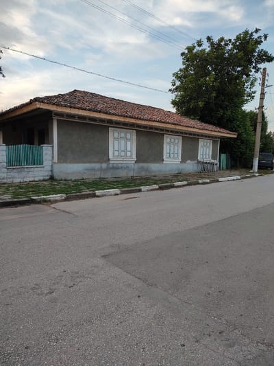 This is a full-frame, eye-level, outdoor shot of a single-story, gray house with a tiled roof. The house is on the left side of the image, viewed from the side, with the front facing away from the viewer. It has three white-framed windows with multiple panes. A green wooden fence stands in front of a section of the house. The house is bordered by a sidewalk with white and black painted curbs, and beyond that is an asphalt road. A large green tree is behind the house on the right, and further back is a utility pole with wires. A dark-colored van is parked on the right side of the road. The sky is partly cloudy with a soft, overcast light. The location is Babuk, Bulgaria.