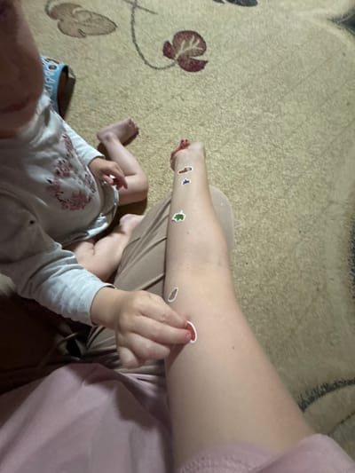 The image shows a close-up of two people's legs and arms on a carpeted floor. One person appears to be an adult, with their lower leg and foot visible, decorated with several small stickers including a red heart, a green leaf, and other small shapes. The other person is a young child, whose legs and arms are also visible. The child is wearing a light-colored long-sleeved shirt with a floral pattern. The child's feet are bare. The background is a textured carpet with a subtle pattern of floral motifs in muted colors. The lighting appears to be indoor, with soft light casting shadows. The mood seems relaxed and playful, suggesting a quiet moment at home. There is no visible text in the image.