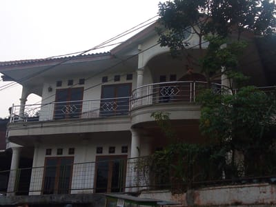 The image shows a multi-story building in Megamendung, Indonesia. The building appears to be a residence, with balconies and windows visible. There are also trees and vegetation surrounding the building, suggesting a semi-rural or suburban setting. The sky is overcast, indicating a cloudy day. The building's architecture features arched windows and a curved balcony, with railings made of metal. The lower levels have iron fences in front of doors and windows. A small stall or kiosk is visible at the bottom of the frame, with the word "BUKA" (which means "open" in Indonesian) printed on it. Electrical wires are strung across the facade of the building.