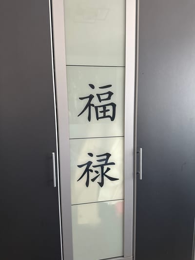 A close-up, eye-level shot captures a section of a modern, dark grey cabinet with a vertical brushed metal handle. The cabinet features a tall, frosted glass panel inset into its center, divided horizontally into two equal sections. Each section displays a large, stylized black Chinese character. The top section features the character "福" (fú), which translates to "fortune" or "blessing." The bottom section displays the character "禄" (lù), meaning "prosperity" or "official salary." The lighting is neutral and even, casting no discernible shadows. The context of Bielefeld, Germany, is not visually present in the image.