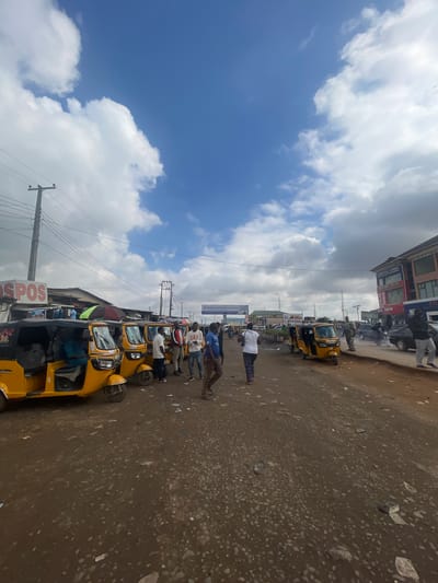 The image depicts a street scene in Ojodu, Nigeria. A line of yellow auto-rickshaws (tuk-tuks) are parked along the roadside. People are standing and interacting near the tuk-tuks, some appearing to be passengers or drivers. The road is unpaved and dusty. Buildings and signage can be seen in the background, including those indicating commercial businesses such as "CHRISVIC LAUNDRY", "MOZIRA GOLD" and "BIG SHOW". A sign with the word "SPOS" is also visible. Power lines are stretched overhead. The sky is partially cloudy.