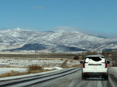 A white SUV drives on a wet road, snow visible on the shoulders. In the distance, layered mountains covered in snow reach towards a clear blue sky. The landscape transitions from the road to a flat expanse with sparse vegetation and patches of snow before rising to the snow-capped mountains. A few buildings are visible in the mid-ground, to the right of the SUV.