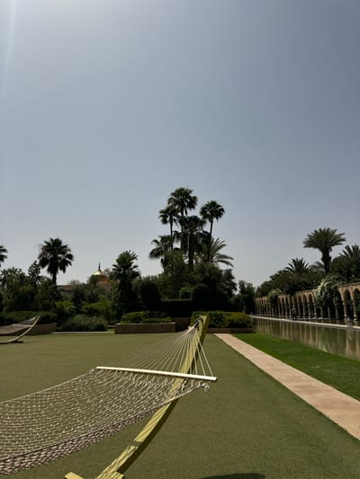 The image shows a hammock in the foreground, set up on a grassy lawn in a manicured garden. The hammock is made of white string and has a wooden frame. Behind the hammock, there is a long, narrow pool of water, with stone arches lining the edge. The water reflects the sky and the surrounding trees. There are several palm trees, lush green bushes, and trees in the background. The sky is a light blue, and the sun is shining. The scene is in Ouled Jelal, Morocco.