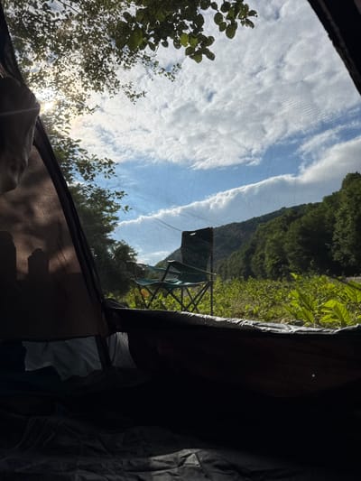 The image shows an outdoor scene viewed from inside a tent. The foreground is dominated by the dark interior of the tent, with fabric textures visible. To the left, a hand is visible, suggesting someone is inside the tent. Sunlight streams through the mesh of the tent opening, illuminating trees with green leaves. Beyond the trees, a steep, lush green mountainside rises under a blue sky with scattered white clouds. A camping chair is visible set up outside the tent, positioned in front of the mountain. The lighting suggests it is daytime, possibly morning or late afternoon due to the angle of the sun. The overall impression is one of a peaceful, natural setting, likely a campsite in a mountainous, vegetated area. The location context provided is Abgara, Abkhazia, which aligns with the mountainous and forested landscape depicted.