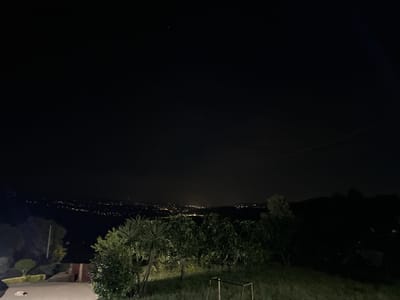 The image depicts a night scene in Celorico de Basto, Portugal. A landscape is visible, with trees and vegetation in the foreground. In the distance, the lights of the city are scattered across the horizon. Above, the night sky is dark, with a few visible stars. A glimpse of a building is noticeable in the left foreground. The image is taken from an elevated viewpoint, possibly a residential area on a hillside.