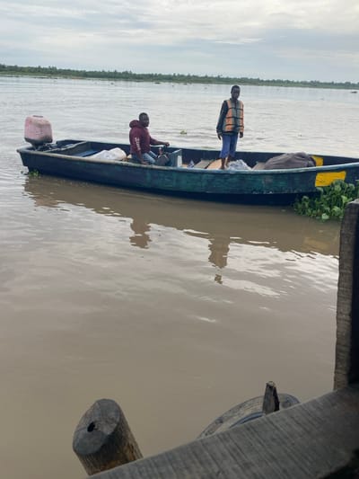The image captures a riverine scene in Akarakumoh, Nigeria, under an overcast sky. Two males occupy a dark green boat, possibly wooden or fiberglass, which is positioned on murky brown water. The boat features a light pink-colored outboard motor, likely covered, mounted on its stern. Yellow markings are visible on the hull near the stern.
One male is seated towards the bow, wearing a maroon hooded sweatshirt with white lettering, blue jeans, and light-colored shoes. His right hand rests on the boat's gunwale. The second male stands bare-footed in the mid-aft section, dressed in a dark long-sleeved shirt, dark blue trousers, and an orange and beige-striped life vest. Both individuals appear to be looking towards the right side of the frame.
Inside the boat, a white sack and a dark grey bundled item are visible. Clumps of green aquatic vegetation are present both in the water adjacent to the boat and partially clinging to its side. The distant riverbank is lined with dense green foliage. In the immediate foreground, a partial wooden structure, possibly part of a dock or pier, is visible, including a dark horizontal beam and a vertical wooden post with a circular cut end and a central dark bore hole. A dark tire, likely serving as a fender, is partly submerged next to this structure.