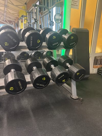 This indoor scene shows a rack of black Tufftech dumbbells, ranging in weight from 22.5KG to 50KG, arranged from light to heavy. The dumbbells are organized on a multi-tiered grey rack. The image includes the text "RE-RACK YOUR WEIGHTS!!!" and "THE TRAINING PARTNER THAT BOOSTS YOUR WORKOUT". The location is a gym in Welshpool, United Kingdom. The environment includes yellow wall decorations.