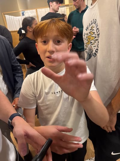 The image depicts an indoor scene in Bergenfield, United States, with multiple individuals. The foreground is dominated by a young male with reddish-brown hair, wearing a white t-shirt with "SONNB LONDON" printed on it. He looks directly at the camera, his mouth slightly ajar, and his right hand is raised in a blurred gesture toward the viewer. To his immediate right, a male in a white t-shirt with a graphic design including "WALLS SINCE" is partially visible. To the left, a person's forearm, adorned with a dark blue watch, holds a dark, elongated object. In the background, a female with dark hair in a ponytail and black attire stands with her hand on her hip, looking right. Further back, a male in a dark baseball cap and another male in a dark green top are visible. The background wall features light-colored panels and white fabric partitions supported by wooden frames.