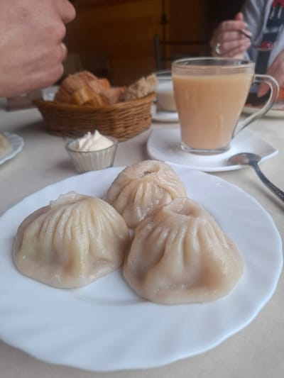 A close-up, eye-level shot shows a white plate with three steamed dumplings arranged in a triangular pattern. The dumplings are pale and have a pleated top. To the left of the plate is a small glass dish containing a dollop of white sauce. Behind the plate, a woven basket contains sliced bread. Further back, a clear glass mug filled with a light brown beverage, likely tea with milk, sits on a saucer. A spoon rests on the saucer. In the background, out of focus, a person's hand is visible, and another person's hands are holding a spoon with a dollop of red food. The scene appears to be set in a cafe or restaurant, possibly in Sukhoy Ruchey, Russia, given the context.
