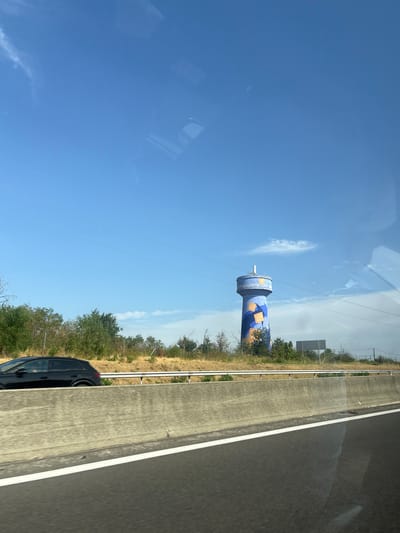 This is a roadside photograph taken from a moving vehicle in the city of Chanas, France. The image captures a bright, sunny day with a clear blue sky and a few wispy clouds. In the distance, a tall, blue water tower stands out, decorated with orange geometric shapes and white text that appears to be a website address. The water tower is surrounded by dry, yellowish grass and scattered green trees. A black car is visible on the left side of the frame, driving on a highway. A concrete barrier runs along the side of the road, separating the highway from the surrounding landscape. The overall impression is one of a typical highway view on a summer day, with the notable presence of the elaborately decorated water tower.