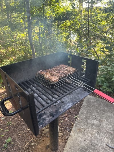 A metal barbecue grill is positioned outdoors in a wooded area, with a concrete slab visible to the right. The grill contains a stainless steel basket filled with meat, cooking over hot coals. Smoke rises from the grill, indicating the cooking process is underway. The surrounding environment consists of trees and foliage, suggesting a park or campsite setting. The image is captured from a medium distance, focusing on the grill and its contents.