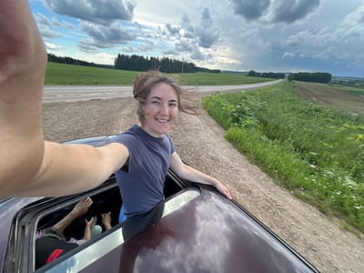 The image captures a young woman leaning out of a car window, with her upper body and head visible. She is smiling and appears to be enjoying herself. The camera is held from her perspective, creating a selfie-style shot. Her left arm is extended outside the car, resting on the vehicle's roof. Inside the car, other individuals are partially visible.
The background shows a rural landscape with green fields and a cloudy sky, suggesting an outdoor setting during the daytime. The car appears to be on a gravel road or a dirt track next to a grassy area. The reflection of the sky and clouds on the car's dark, glossy surface is noticeable.
The image is oriented sideways, with the subjects appearing upside down. The clothing of the woman is a plain dark t-shirt. A red tag with the word "HUGO" is visible on a black item of clothing inside the car. The overall mood seems lighthearted and adventurous, possibly indicating a road trip or a scenic drive. The location context provided is Верх-Рождество, Russia.