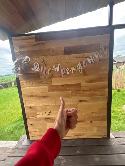 A wooden panel with a banner reading "She said yassss" in cursive font hangs in front of a blurred outdoor scene in Lahojsk selsaviet, Belarus. A partially visible balloon with interior decorations is situated at the bottom of the banner. A human arm wearing a red sweater is extended towards the wooden panel with the hand forming a thumbs-up gesture. The panel is framed in dark wood or metal and appears to be part of an outdoor structure or backdrop. The background shows green grass and trees, as well as a fence and a structure in the distance.