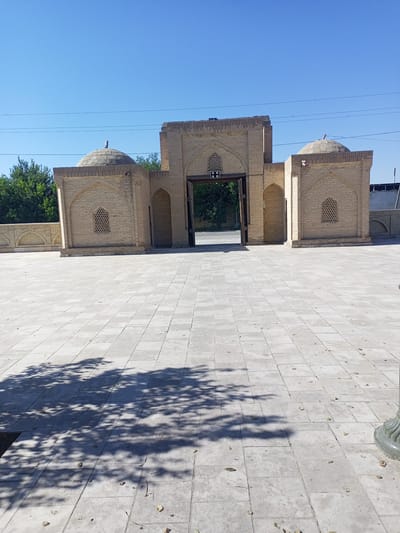 This is a medium shot, outdoor, eye-level photograph of the main entrance to the Chor-Bakr necropolis in the city of Shakhrisabz, Uzbekistan. The entrance is a large, arched structure made of light-colored brick, with two smaller domed buildings on either side. The ground in front of the entrance is paved with large, square tiles. The sky is clear and blue, and there are trees in the background. Shadows of trees are cast on the tiled ground. The overall scene is one of historical architecture and serene public space.