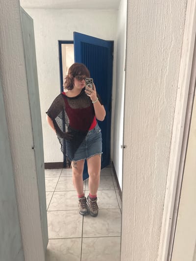 A mirror selfie shows a person with short brown hair wearing a red top, a black sheer shirt, and a denim skirt. Pink socks peek out of hiking boots. A turquoise bracelet is visible on the left wrist. The person is holding a smartphone. The background features a white textured wall above and tiled flooring below, divided by a blue door and frame.