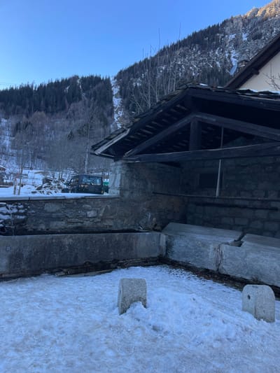 This image captures a serene winter landscape in a mountainous region, consistent with the Courmayeur, Italy, context. The scene is dominated by a traditional stone building with a dark wooden roof structure, visible on the right side of the frame. Its sturdy stone walls and exposed timber beams suggest classic alpine architecture.

The foreground and mid-ground are covered in a blanket of snow, with some patches of bare earth visible. Two distinctive, rectangular stone blocks, possibly bollards or markers, are partially submerged in the snow close to the viewer. A longer, lower stone structure runs parallel to the building's base, forming what appears to be a pathway or a low retaining wall.

Further back, near the building, a dark-colored vehicle, resembling an excavator or small tractor, is parked on the snowy ground. Beyond the building and vehicle, a rugged mountain slope rises majestically, densely covered with a mix of bare deciduous trees and dark green coniferous trees, indicating a forest. The upper portion of the scene is dominated by a clear, vibrant blue sky.

The time of day appears to be daytime, with strong, clear light suggesting a sunny winter day. There is no visible activity taking place, contributing to a peaceful and quiet atmosphere. No people are visible, and there is no discernible text in the image. The overall impression is one of tranquil natural beauty combined with traditional mountain habitation.