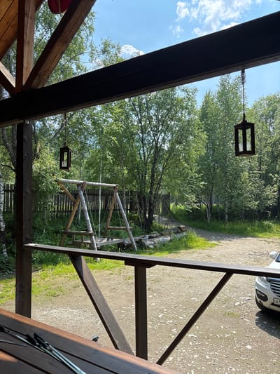 The image captures a view from a wooden porch in Bol'shaya Rechka, Russia. In the foreground, a wooden table is partially visible with skewers laid out on it. A dark brown wooden railing separates the porch from the outdoor area. Beyond the railing, a gravel pathway leads into a lush green area dominated by trees. A wooden swing set stands prominently in the middle ground, its rustic construction blending with the natural surroundings. A wooden fence runs along the background, and further back, a white car is parked. The sky above is a clear blue with scattered white clouds. Two dark, square lanterns are suspended from the porch roof, one on the left and another on the right, adding to the rustic ambiance of the scene.