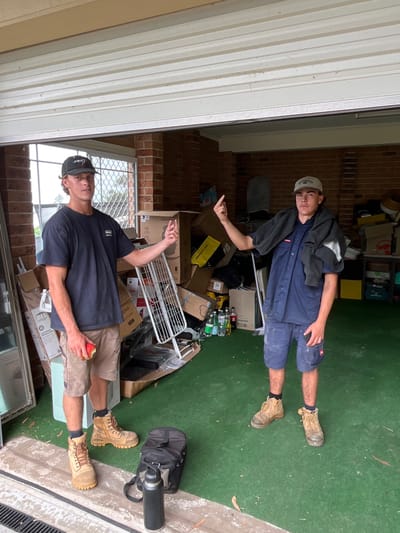 The image depicts two young adult males standing within what appears to be a cluttered residential garage in Wombarra, Australia. The floor is covered by a green mat, transitioning to a concrete threshold in the foreground. The background features a brick wall, a window with security bars on the left, and numerous stacked cardboard boxes containing various household items. A white roller garage door is partially open at the top of the frame.

The male on the left wears a dark baseball cap, a dark t-shirt, khaki shorts, and tan work boots. He holds a partially consumed red apple in his left hand. His right arm is raised, with his index finger extended upwards.

The male on the right wears a light baseball cap, a dark blue short-sleeved work shirt, dark blue cargo shorts, and tan work boots. A dark garment is draped over his left shoulder. His right arm is raised, with his middle finger extended upwards.

Between the two subjects in the foreground, a black backpack and a black insulated water bottle rest on the concrete. A white metal drying rack is visible among the boxes behind the subjects.