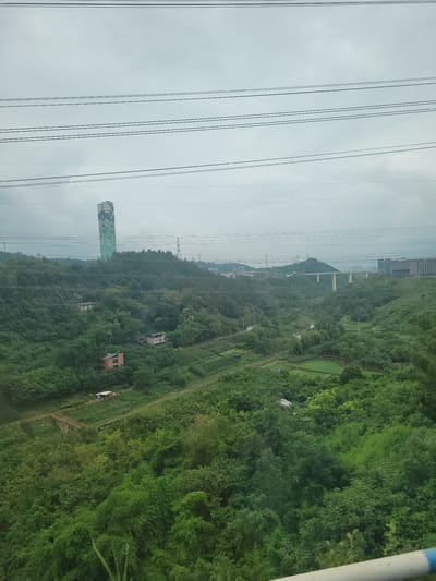 This image depicts a scenic view from a cable car traversing over a lush, green landscape in Yufengshan, China. The primary subjects are the dense forest covering rolling hills, interspersed with a few buildings and infrastructure elements. In the foreground, multiple parallel power lines stretch across the frame, indicating the presence of electrical infrastructure.

Further into the landscape, a distinctive tall, green and white striped tower with some text visible on its side stands out against the trees. Several small, brick-colored buildings, appearing to be houses, are nestled within the foliage. A white bridge or elevated structure is visible in the distance, spanning across a hillside. The overall scene is one of natural beauty with a touch of human development.

The activity taking place is the operation of a cable car system, suggested by the perspective and the view from what appears to be inside a car. The time of day seems to be daytime, given the natural light illuminating the scene. The weather appears to be overcast or hazy, as indicated by the muted sky. The location is identified as Yufengshan, China. No specific emotions are conveyed, and the notable details include the power lines, the distinct striped tower with text, the small houses, and the bridge, all set against a backdrop of dense greenery.