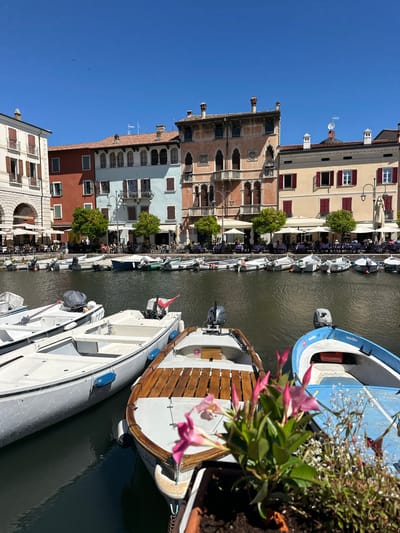 This media file depicts a sun-drenched canal scene in Desenzano del Garda, Italy. The perspective is from above, looking down onto a waterway lined with colorful buildings and several boats. The buildings feature Italian architectural elements, with varied facades in shades of cream, ochre, light blue, and terracotta, adorned with red-shuttered windows and intricate detailing.

A series of boats are moored along the canal. These include a prominent blue and white boat in the foreground, a classic wooden-decked boat, and several smaller white vessels. Each boat has an outboard motor. The canal itself is a deep, still green, reflecting the bright blue sky and the surrounding structures.

Along the canal's edge, a strip of cafes or restaurants is visible, with tables and chairs set up outdoors under awnings and umbrellas. Scattered trees with lush green foliage add a touch of nature to the urban setting. The presence of people can be seen at the outdoor seating areas, suggesting a leisurely daytime activity.

The weather appears to be clear and sunny, indicated by the bright sky and strong light casting shadows. The overall mood is tranquil and picturesque, capturing the essence of a charming Italian waterside town on a pleasant day. There is a visible number "41" on one of the boats. A cluster of pink flowers with green leaves is prominently featured in the foreground, adding a splash of color and detail.