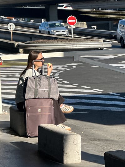 The image, oriented correctly, depicts a person sitting on a concrete barrier or bollard in an urban outdoor setting on a bright, sunny day, likely in Tremblay-en-France, France. Strong sunlight casts sharp, geometric shadows from nearby structures onto the asphalt ground, which features white painted markings, including stripes and abstract patterns.
The primary subject is a person, likely female, with long dark hair, wearing sunglasses on her head. She is dressed in dark trousers and a light-colored top, and wears white sneakers with three dark stripes. She is holding a piece of yellow fruit or food in her right hand. Beside her are two pieces of luggage: a grey duffel bag with the brand "Avène" visible on it, and a brown wheeled suitcase with a subtle logo that is difficult to decipher.
In the background, a road with white lane markings is visible, along with multiple vehicles, including a white car, a silver van (with part of a French license plate "GM-187-..." visible), and another dark vehicle. Guardrails and concrete barriers separate different levels or sections of the road infrastructure, suggesting an elevated road or bridge, possibly near an airport given the luggage and location context of Tremblay-en-France (home to Charles de Gaulle Airport). A prominent red circular "Do Not Enter" sign with a white horizontal bar is clearly visible on a pole in the mid-ground. The person appears to be resting or waiting, possibly during travel.