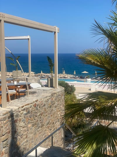This is a medium-shot, eye-level, exterior photograph taken at a resort in Panormos, Greece. The scene depicts a stone wall in the foreground, leading down a set of stairs with a stainless steel handrail. To the right, palm fronds are visible. Beyond the wall, a swimming pool with lounge chairs and umbrellas is situated, overlooking the bright blue Aegean Sea. The sky is clear and blue. The overall ambiance is tranquil and idyllic.