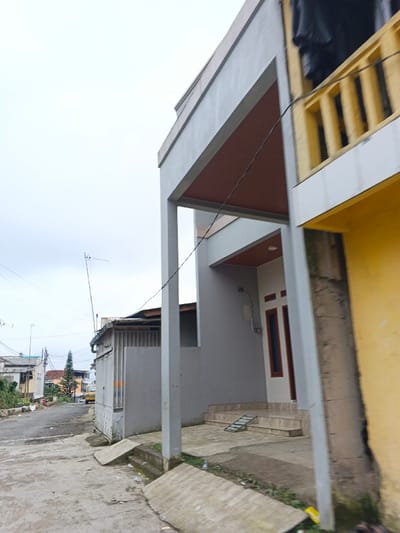 This image depicts a street scene in Kabiufa, Papua New Guinea, under an overcast sky. The foreground features a concrete ground surface with two concrete ramps leading to an elevated concrete patio. To the right, a two-story light gray building with a flat roof and a recessed entrance is visible. This entrance is supported by two square pillars, with the underside of the overhang and a section of the facade rendered in reddish-brown. A dark brown-framed door, featuring two narrow vertical windows, is situated within this recess, accessed by a series of concrete steps. A white utility box is mounted on the wall adjacent to the door. To the immediate right, a yellow-painted building is partially observed, displaying an upper-level balcony with a yellow balustrade and a black tarp draped over its top edge. To the left of the light gray building, a smaller, light-colored structure with vertical metal grating is present. A paved road curves into the background on the left, revealing additional residential buildings and a slender conifer tree in the distance. Overhead utility wires are visible, and a television antenna is mounted on a pole to the left of the central gray building. No individuals or vehicles are present.