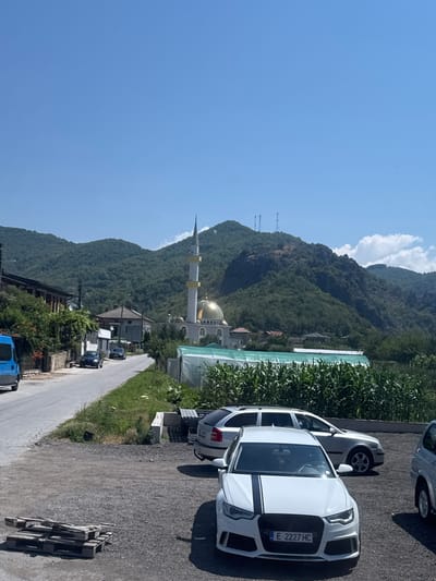The image depicts a sunny day in a village setting, likely in Gospodintsi, Bulgaria, given the visible mosque. In the foreground, a gravel lot contains several parked cars, including a white Audi with black racing stripes on its hood, a silver station wagon, and a silver SUV. Wooden pallets are stacked on the left side of the lot. A paved road runs along the left side of the image, with a blue van parked near a row of buildings on the left. Further down the road, two dark-colored cars are visible. In the midground, a white mosque with a golden dome and a tall minaret stands prominently. A greenhouse structure with a white covering is situated to the right of the mosque. Beyond the mosque and greenhouse, a densely forested mountain range forms the backdrop, with clear blue skies overhead, dotted with a few white clouds. The overall scene suggests a peaceful rural community with a mix of residential buildings, religious architecture, and agricultural elements.