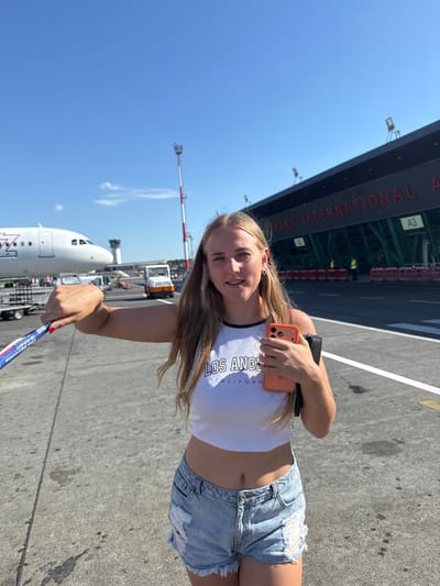 A young woman with blonde hair is posing for a photo at an airport, with an airplane visible in the background. She is wearing a white crop top with "LOS ANGELES CALIFORNIA" printed on it and ripped denim shorts. She is holding an orange smartphone in her right hand and a lanyard with text in her left hand. The airport terminal is visible in the background with large red lettering. The sky is clear and blue, suggesting a sunny day. The scene appears to be at an airport apron or tarmac. The woman is smiling at the camera.