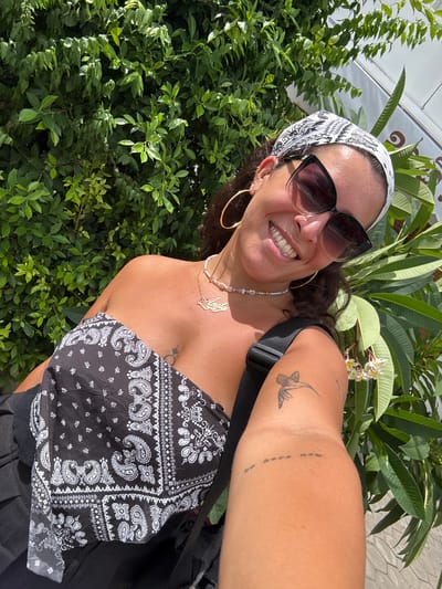 A woman takes a selfie outdoors with a lush green bush behind her. She is wearing a black and white paisley bandana as a headscarf, large sunglasses, and a black strapless top with a paisley print. She has large hoop earrings and a delicate necklace with pearls and a gold nameplate. A tattoo of a hummingbird is visible on her right arm, along with a small text tattoo. The woman is smiling broadly and tilting her head. The environment appears to be sunny and tropical. The photographer is positioned to capture the woman from the chest up, with her right arm extended to take the photo. The scene is devoid of any specific visual cues that would indicate it is located in Kampot, Cambodia.