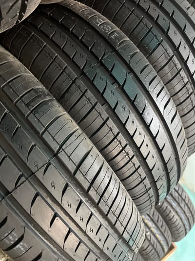 A close-up, angled, vertical shot displays a stack of several new, black car tires. The tires are arranged in a staggered fashion, with their tread patterns clearly visible. The texture of the rubber is detailed, showing small circular indentations and grooved channels designed for traction. The lighting highlights the contours of the tread, creating a play of light and shadow. There are no people or any other discernible objects in the frame besides the tires. The background is indistinct and appears to be a plain, light-colored surface, possibly a wall or a display area. The image does not provide any specific geographical or contextual information that would allow for identification of a location in Furmanov, Russia.