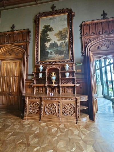 The image depicts a room interior, likely in the Vorontsov Palace, located in the city of Алупкинский городской совет, Ukraine. The scene includes a large painting on the wall, depicting a landscape. Below it is a carved wooden cabinet with open shelves displaying decorative vases. Carved wooden paneling is present on the walls and below the cabinet. The floor is parquet. The area above the cabinet is a glazed ceiling, letting in natural light. A sign with "ВЫХОД" (Exit) in Russian is visible.