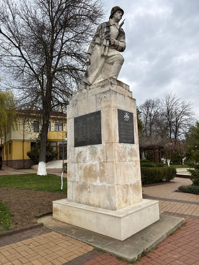 The image captures a monument in Chavdar, Bulgaria, featuring a stone statue of a soldier set upon a large, multi-tiered concrete or stone pedestal, under an overcast sky. The statue depicts a male soldier in a helmet and uniform, dynamically posed as if leaning back or resting, with a rifle or machine gun held across his chest. He also appears to have binoculars or grenades strapped to his waist, and his gaze is directed upwards and slightly to the left.
Two dark rectangular plaques are affixed to the front of the pedestal. The upper plaque features a star-like emblem with rays and partially legible text, prominently displaying "ЗА РОДИНАТА" (For the Motherland). The lower plaque contains a list of names, likely of fallen soldiers, with "БЕЛИШКИ" and "СТОЯН" being visible among others.
The monument is surrounded by bare-branched deciduous trees, suggesting late winter or early spring. The lower portion of one tree trunk near the monument is painted white. In the background, a building with yellow walls and brown window frames is visible. The ground features sections of green grass, some areas of exposed dirt, and paved walkways constructed from reddish and brown interlocking bricks, alongside some grey concrete slabs. The sky is uniformly grey and overcast, creating a muted lighting and a somber atmosphere. No specific activity is taking place, presenting a static scene of remembrance.
