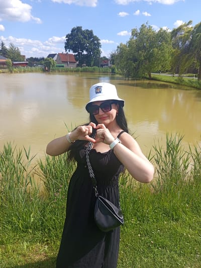 An adult female stands on a grassy bank beside a murky, yellowish-brown pond under a bright blue sky with scattered cumulus clouds, in the city of Ptice, Czechia. She is positioned centrally in the frame, facing forward, forming a heart shape with her hands in front of her chest.

The woman wears a white bucket hat with the text "CAPYBARA BRUH" in blue and a graphic of a capybara. She has dark, oversized sunglasses, small red earrings, and long, dark hair visible beneath the hat. Her attire includes a black, off-the-shoulder dress or top, and a black crossbody bag with a white-striped strap and silver hardware. On her left wrist are multiple silver-toned bracelets, and on her right wrist is a white smartwatch or fitness tracker.

The immediate foreground consists of lush green grass, transitioning to taller, reedy vegetation along the water's edge. The pond's surface reflects the sky and distant trees. In the background, across the pond, a rural or village scene unfolds. To the left, several residential buildings with pitched roofs are visible amidst dense green trees and shrubs, including a large, prominent deciduous tree. A green vehicle, possibly a bus, is partially visible near the houses. To the right, beyond the pond, a paved road with white edge markings runs parallel to the water, bordered by more trees, some appearing wind-swept. The lighting indicates a sunny day.