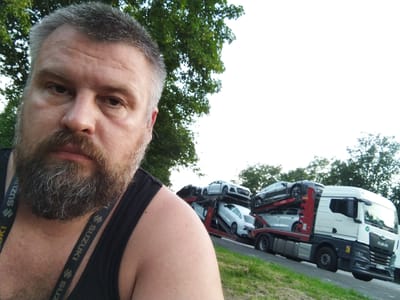 A selfie is taken by a man with a beard, wearing a black tank top with Suzuki lanyards. The man is positioned on the left side of the frame, with his face and upper torso filling a significant portion of the left side. In the background, on the right side of the frame, a car carrier truck is loaded with several cars. The truck is white and grey, with the "MAN" logo visible on the front. The cars on the carrier are a mix of white and dark colors, some appearing to be sedans or coupes. The truck is on a paved surface, with a grassy embankment and trees on the right side of the image. The sky is overcast. The image appears to have been taken outdoors during the day. The location in the city of Loxstedt, Germany, is indicated by the presence of a white MAN truck, which are commonly used in the region, and the overall rural setting often found outside of more densely populated areas in Germany.