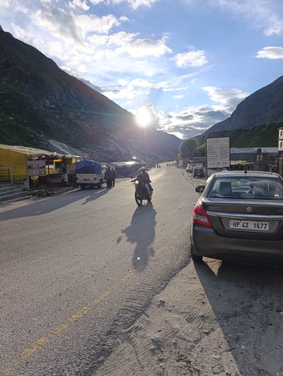 In Damphug, India, a sun-drenched asphalt road winds through a valley flanked by steep, rocky mountains. The sun, low in the sky, casts long shadows and illuminates scattered clouds. Along the left side of the road, temporary stalls with yellow awnings display goods, including food items labeled "MAYA DHABA". A white pickup truck with a blue tarp covers its load is parked. Two men stand near the truck. Further down the road, a motorcycle with a rider wearing a helmet and a face mask is in motion. On the right side of the road, a brown Maruti Suzuki sedan is parked, its license plate reading "HP 42 1677". The road surface is a mix of asphalt and gravel, with tire tracks visible in the sandy shoulder. A large informational sign stands near the parked car. The overall scene depicts a roadside stop in a mountainous region.