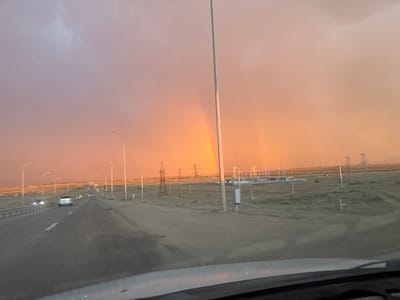 The media file depicts a scenic view of a highway in Satbayev, Kazakhstan, during what appears to be sunset or sunrise. The sky is a prominent feature, displaying a gradient of colors from a dusky purple at the top to a vibrant orange and yellow near the horizon, suggesting dramatic lighting conditions. A faint double rainbow is visible in the distance, adding to the atmospheric beauty of the scene.
The foreground is dominated by the road, with a white vehicle in the center lane. The road is bordered by a guardrail on the left and a dusty, open landscape on the right. Numerous streetlights line the highway, their lamps casting a faint glow. In the middle ground and background, power lines and transmission towers are scattered across the plain, indicating an industrial or utility area. In the far distance, wind turbines are visible, further contributing to the impression of an open, somewhat developed landscape. The overall mood of the image is serene and awe-inspiring, likely evoking a sense of wonder due to the natural phenomenon of the rainbow and the colorful sky. The time of day is clearly indicated by the lighting and the sky's colors, pointing to either dawn or dusk. The weather appears to be clear, with the exception of the rain that caused the rainbow. There is no visible text in the media file.