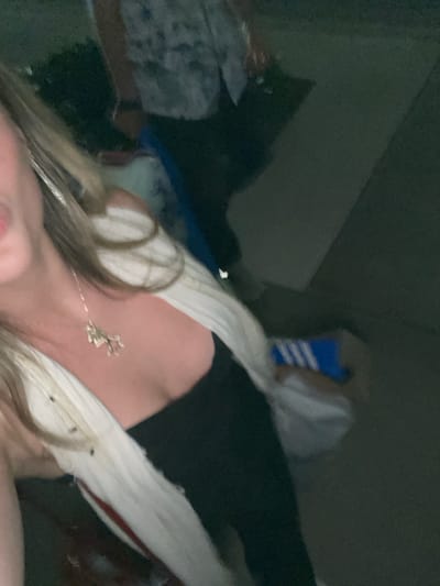 The image captures a close-up, angled shot of a person, likely taken from a low perspective looking upwards. The overall scene is dimly lit, suggesting an indoor setting with low ambient light, possibly at night.
The central focus is on the upper body of the person. Long, light-colored hair, appearing to be blonde or light brown, cascades across the bottom and left side of the frame, partially obscuring the person's chest. A delicate silver-toned necklace with an intricate, small charm (resembling a leafy design or abstract shape) rests on the person's chest.
The person is wearing a light-colored garment, possibly white or cream, which appears to be made of a ribbed or textured fabric, draped over the shoulders and chest. A darker piece of clothing, possibly black, is visible underneath the light garment on the right side of the frame.
The background is very dark and indistinct, making it difficult to discern specific objects or details. However, vague shapes suggest the presence of furniture or other objects in a cluttered or dimly lit room. On the upper right, there's a faint hint of a blue object with white stripes, possibly a bag or box, but its exact nature is unclear due to the low light and blur.
The image conveys a casual and relaxed atmosphere, with the person appearing to be in a reclining or lying down position. There is no visible text in the image.