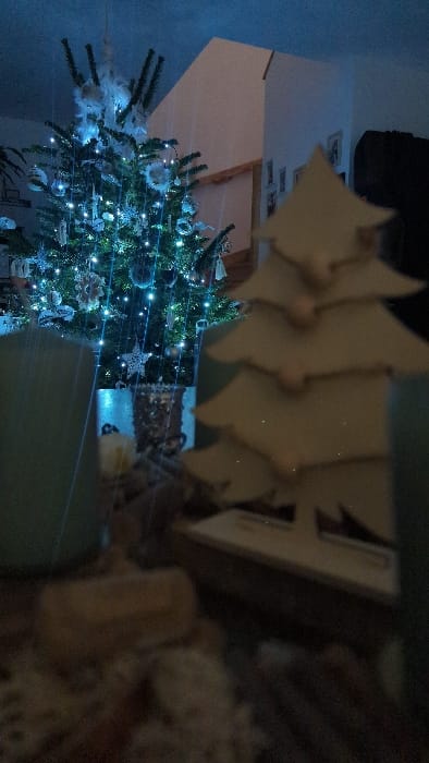 A decorated Christmas tree is seen indoors, adorned with lights and various ornaments. A wooden Christmas tree stands nearby, and a blurred photograph of people sits to the right of the wooden tree. The wall has a light blue hue.
