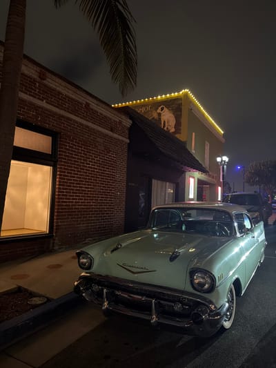 This image depicts a nighttime street scene in El Segundo, United States of America. In the foreground, a light-green 1957 Chevrolet Bel Air classic car is parked curbside, facing right. The vehicle displays prominent chrome bumpers, grille, headlight bezels, hood ornaments, and whitewall tires.
To the left of the vehicle, a red brick building features a large, brightly illuminated display window revealing an empty, white-walled interior. A palm tree trunk partially obstructs the left side of this building, with its fronds visible in the upper left. Adjacent to the brick building is a light green structure with a dark, sloped roof covering an entrance. The upper facade of this building features a mural depicting a white dog listening to a gramophone, with the word "Victor" partially visible. A string of warm yellow incandescent lights adorns the building's roofline. Below the mural, a sign identifies the location as "OLD TOWN MUSIC HALL," flanked by two vertical, illuminated display cases at the entrance.
A dark SUV or similar vehicle is parked further down the street behind the classic car. Streetlights, including one with a double lamp fixture and another emitting a blue hue in the distance, provide additional illumination. The sky is dark, indicating night. No individuals are visible in the scene.